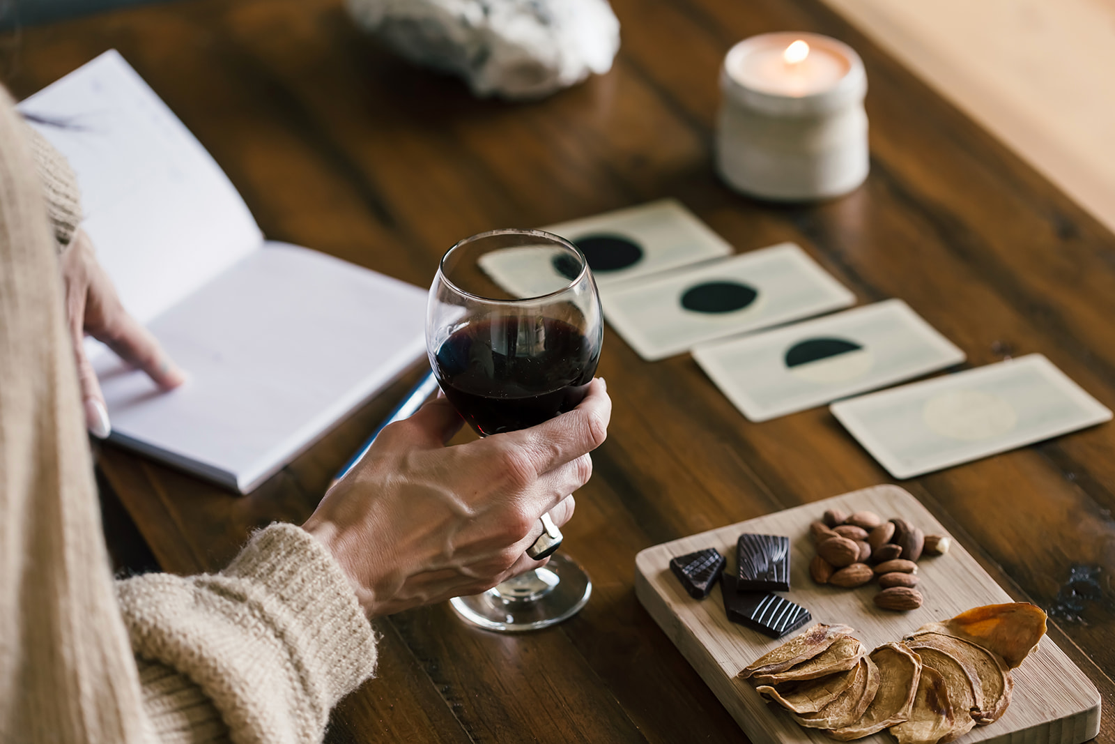 Flatlay of a cozy tarot ritual setup with a woman holding a glass of red wine, oracle cards on a wooden table, a candle, notebook, and a snack board with chocolate and almonds.