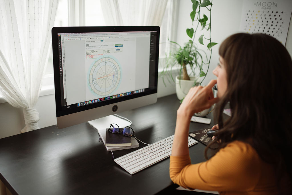 Moonstone founder Miranda analyzing an astrological birth chart on a desktop computer using astrology software, with notebooks and glasses on the desk—astrology-based brand strategy workspace.